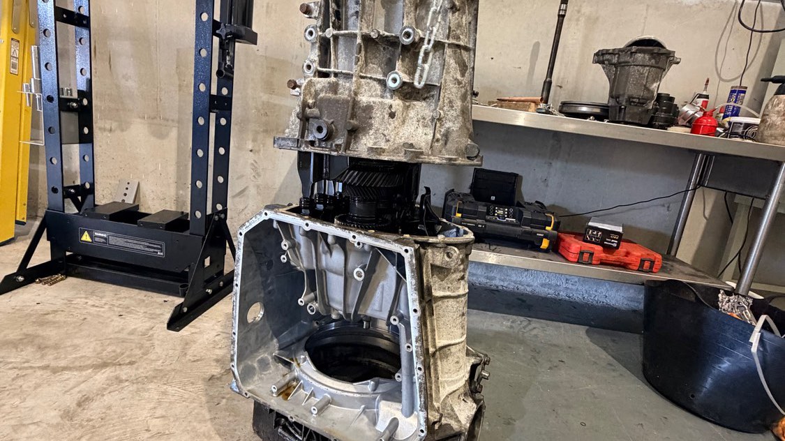 A DL501 0B5 seven-speed S tronic gearbox split open on a chain hoist, the front case lifted clear of the rear housing with the clutch pack and gear train exposed, in the Leek workshop