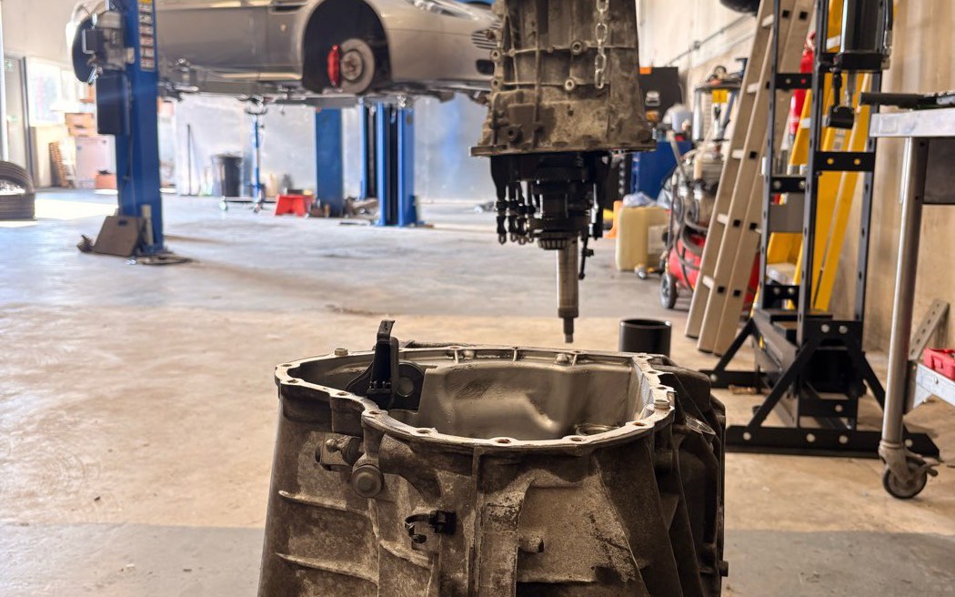 A gearbox split open on the hoist in the foreground of the workshop, with a car raised on the two-post ramp behind it